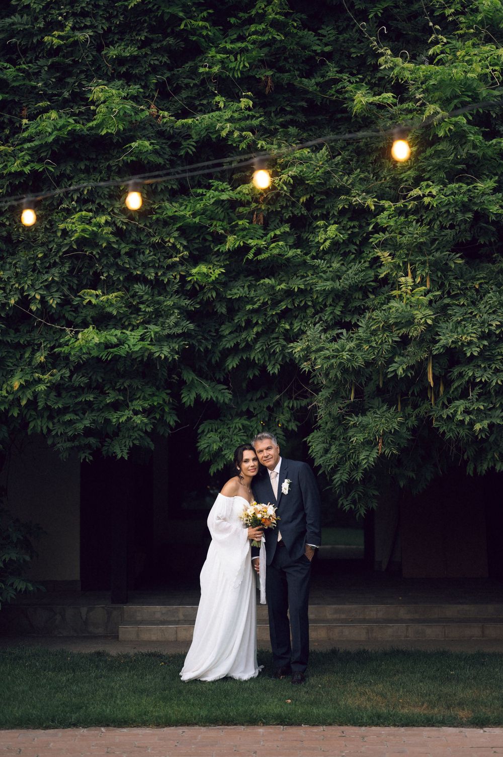 Bride and groom posing together outdoors under string lights with lush green ivy wall backdrop on their wedding day.