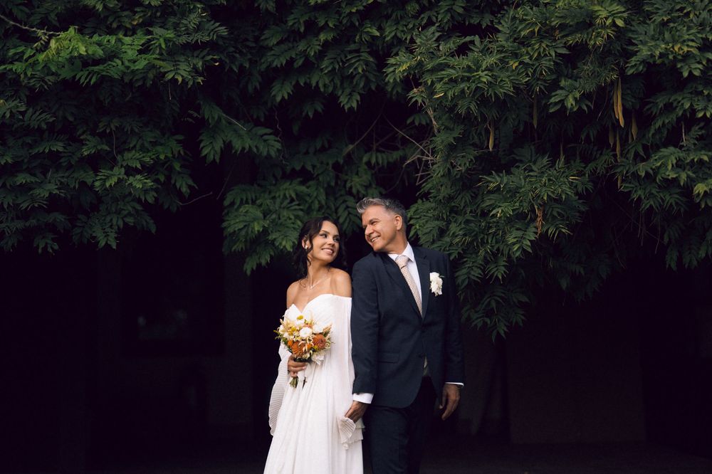 A bride and groom pose together outdoors in front of lush green trees, she holds a yellow bouquet in a white gown.