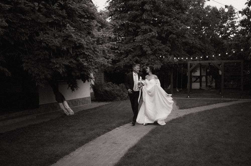 Black and white photo of a bride and groom dancing outdoors on a garden path with string lights and trees.