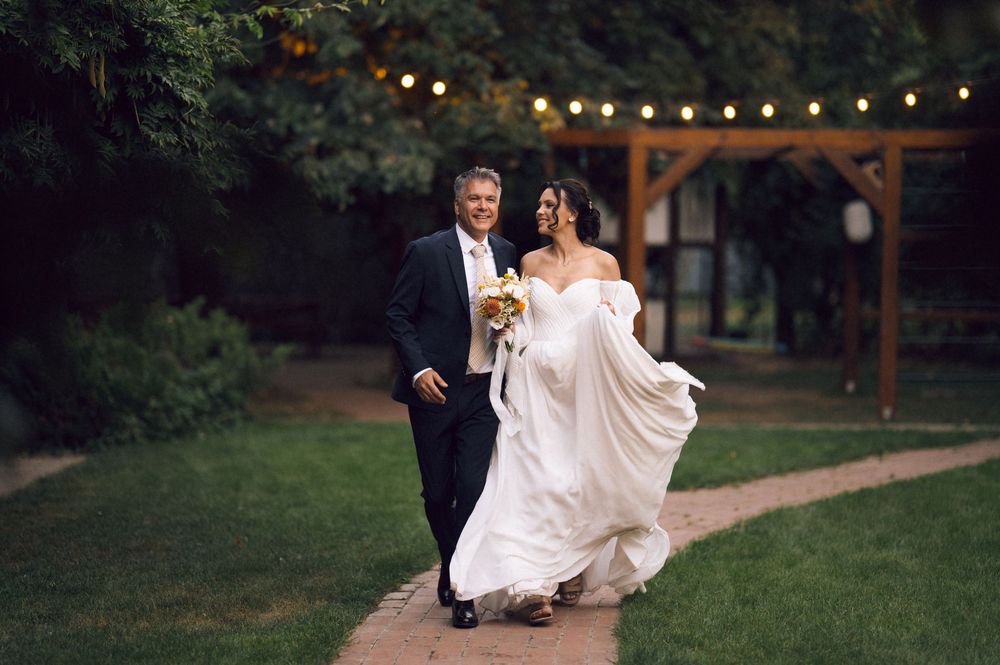 A bride and groom walk hand-in-hand through a garden venue with string lights at their outdoor evening wedding.