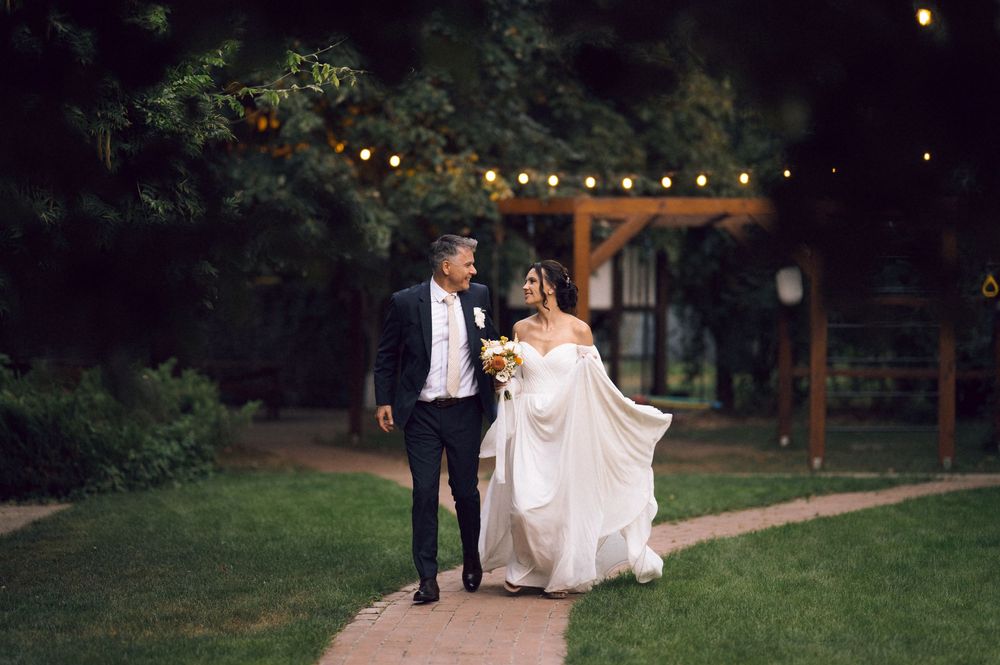 Bride and groom walk hand-in-hand through a garden at dusk with string lights glowing in the background.