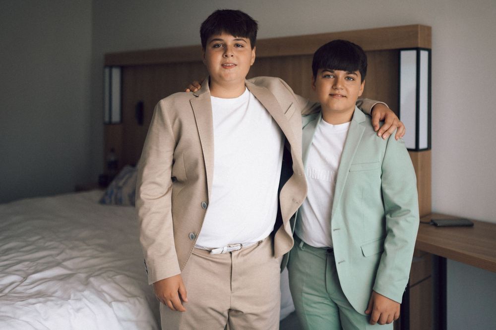 Two boys in blazers pose together in a bedroom, one in beige and one in mint green suit jackets.