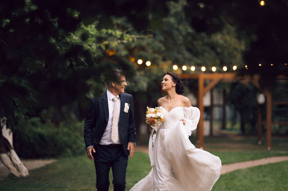 Bride in white off-shoulder gown holding bouquet walks with groom in dark suit at outdoor evening wedding reception.