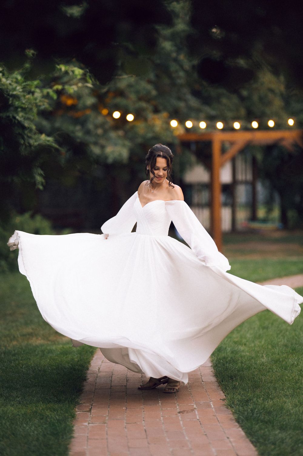 Bride in flowing white off-shoulder gown twirling on brick path in lush garden with string lights.