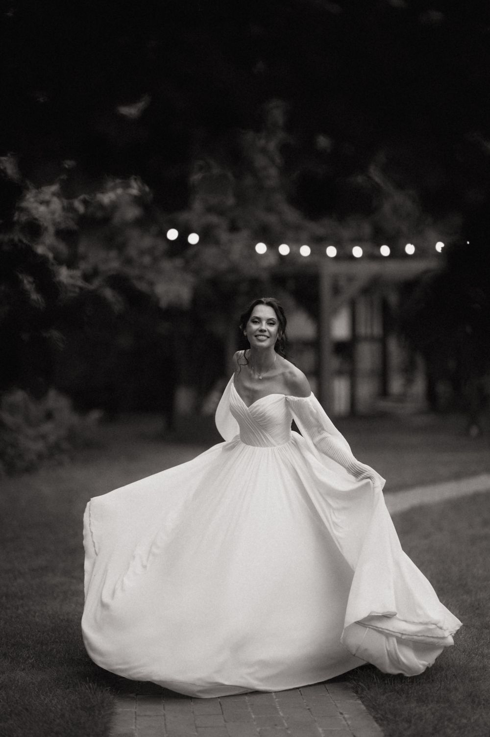 Smiling bride twirls in elegant off-shoulder ballgown outdoors at night with string lights in black and white photo.