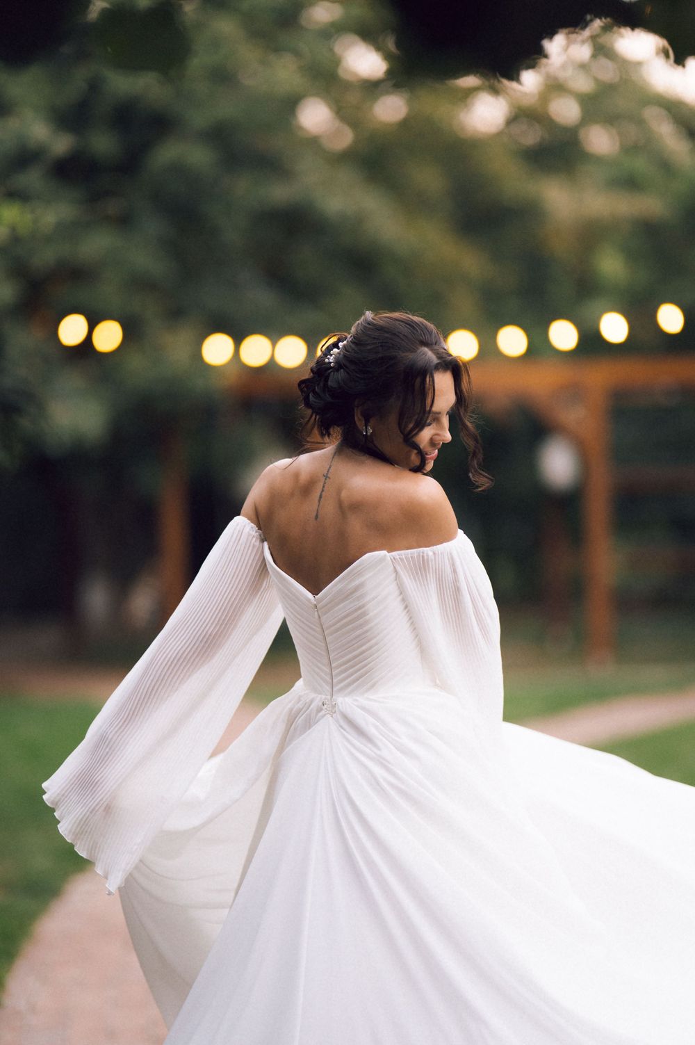 Bride in off-shoulder white wedding gown twirling outdoors with string lights and lush greenery in background.