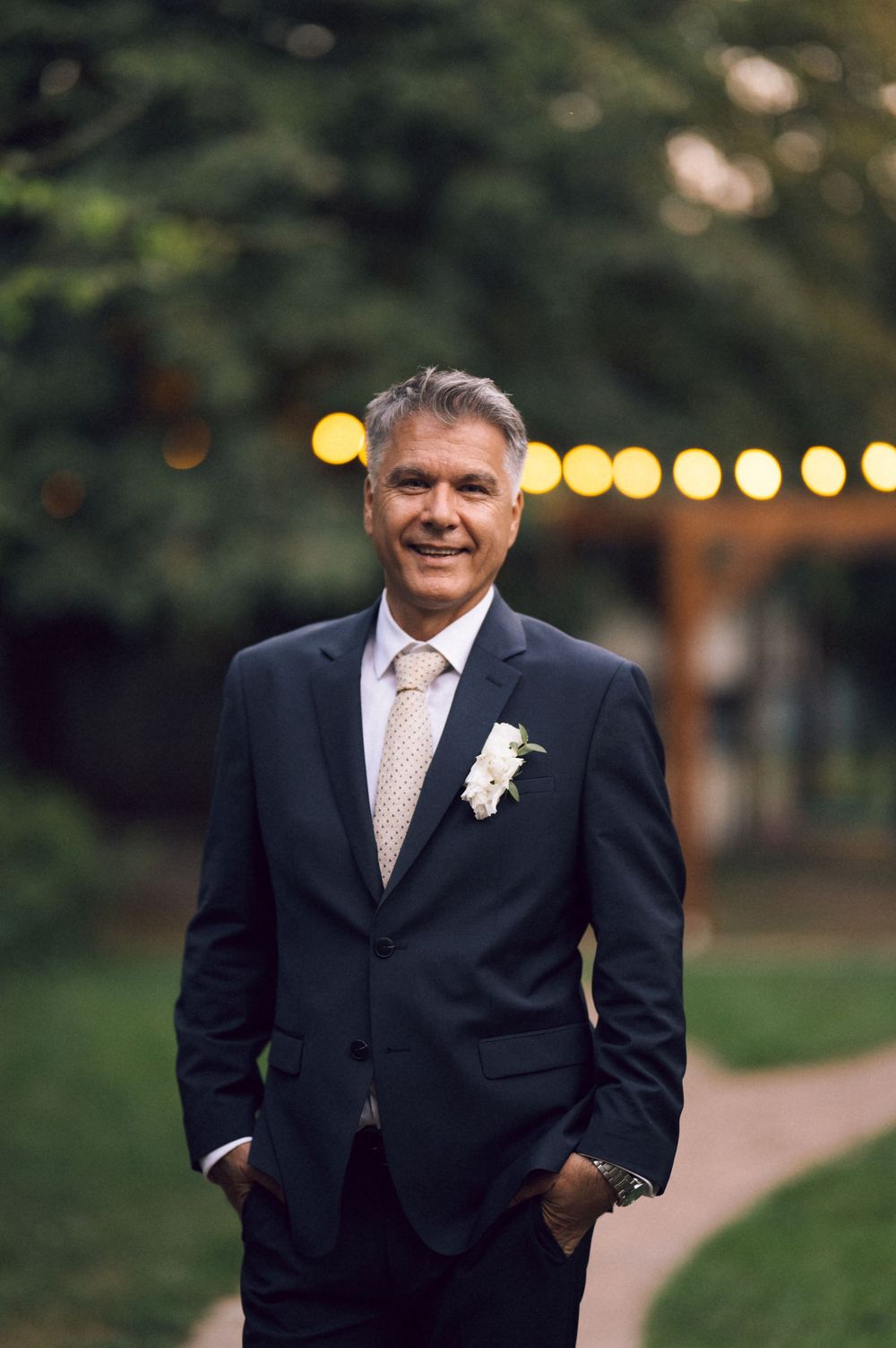 Smiling middle-aged man in navy suit with boutonniere stands outdoors at wedding venue with string lights.