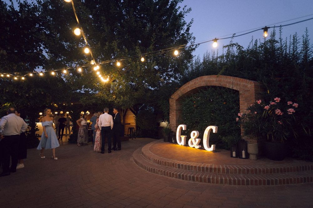 Outdoor wedding reception at dusk with string lights, brick archway, and illuminated G&C monogram letters.