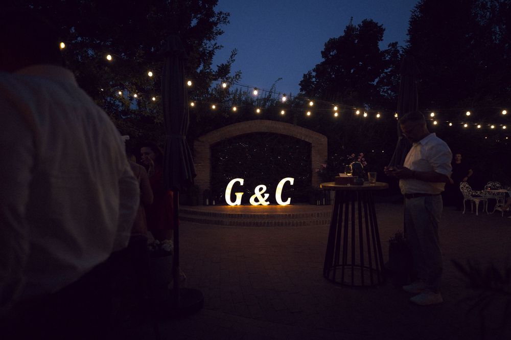 Outdoor evening wedding reception with glowing G&C monogram letters and string lights in a garden venue.