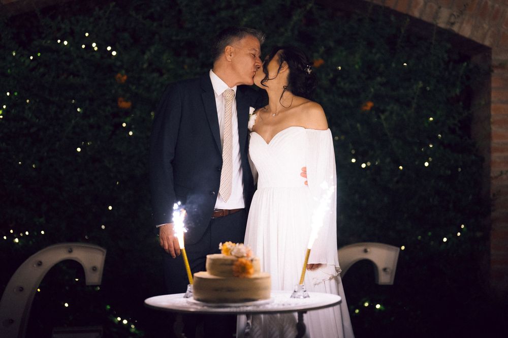 Couple sharing a romantic kiss at their wedding reception at night, with a decorated cake and sparkler on the table.