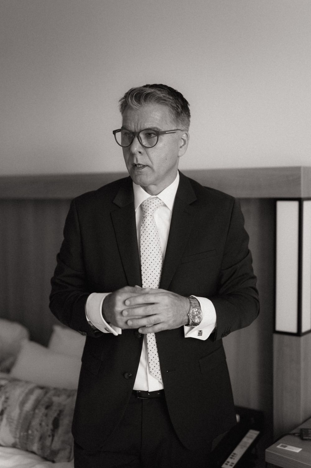 Black and white portrait of a middle-aged man in a dark suit and dotted tie, wearing glasses, standing in a hotel room.