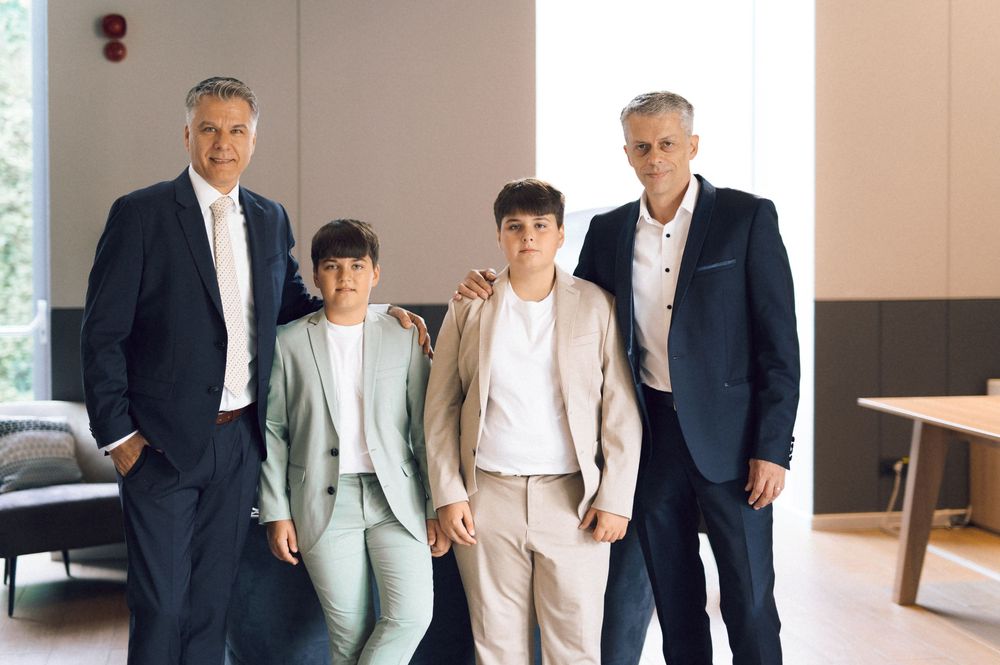 Two men in suits pose with two children in formal attire in a modern interior setting.