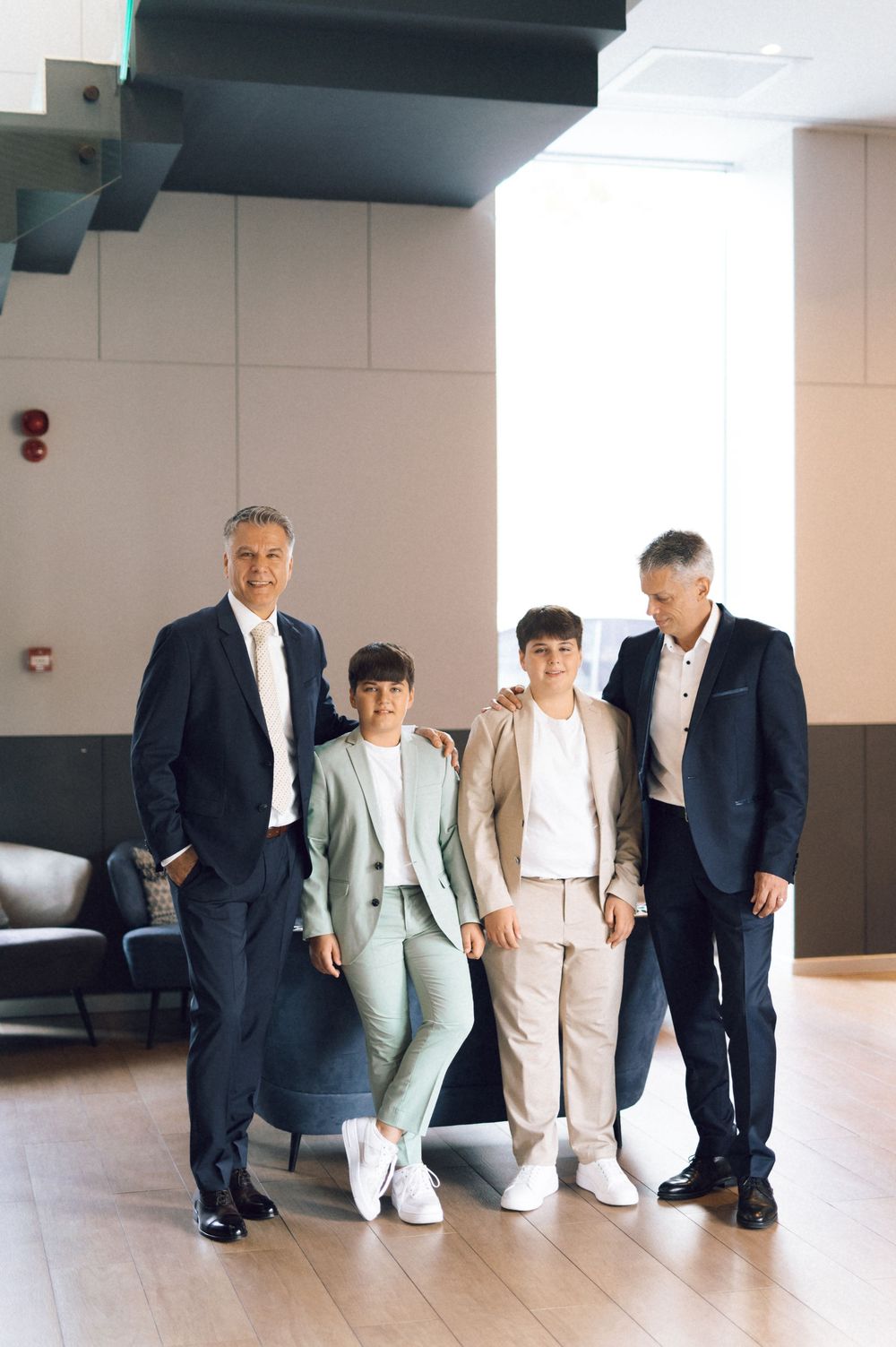 Four people in formal suits posing together in a modern building interior with wooden floors.