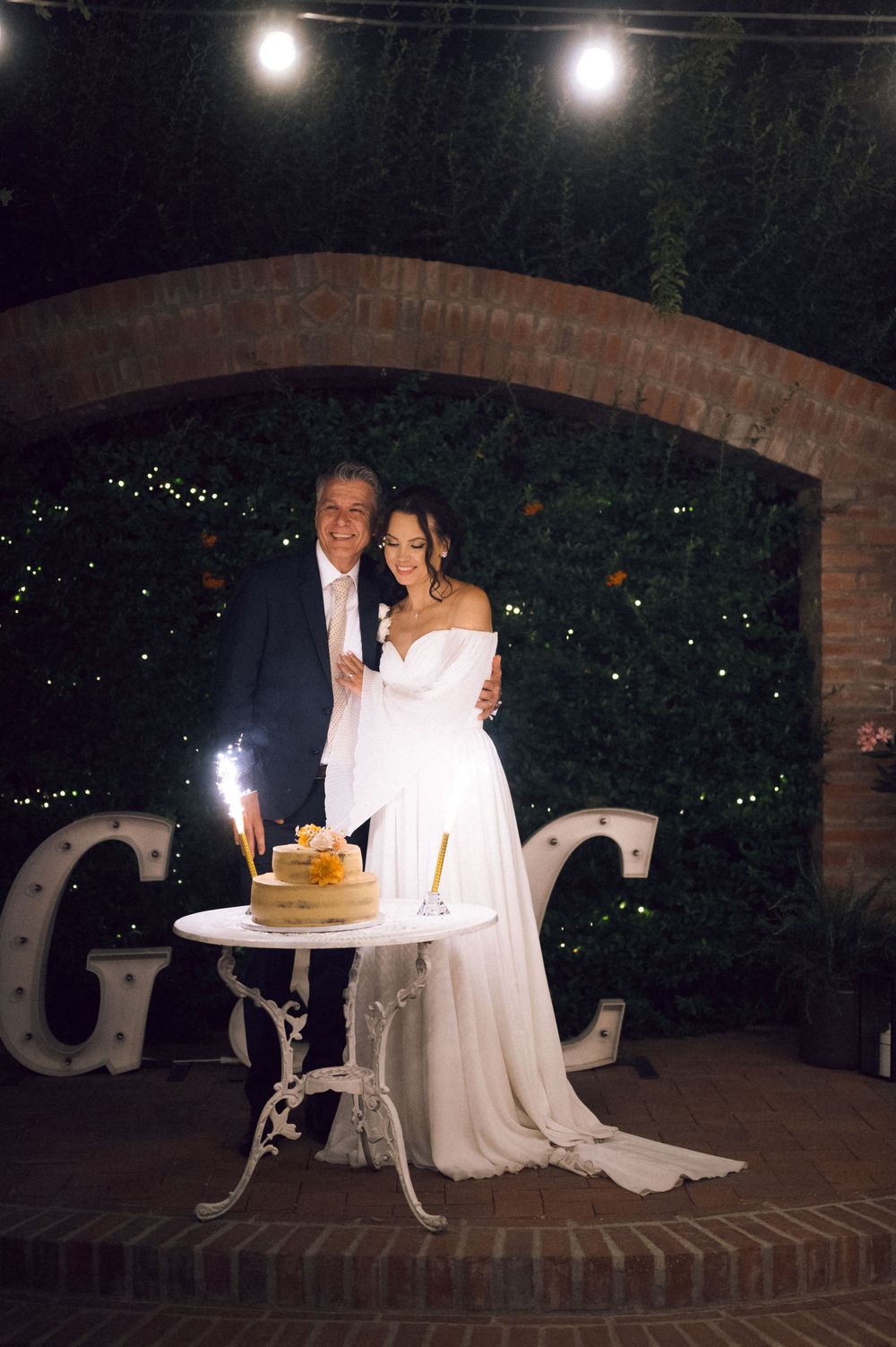 Bride and groom cutting wedding cake outdoors at night under brick arch with sparkler and illuminated letter decorations.