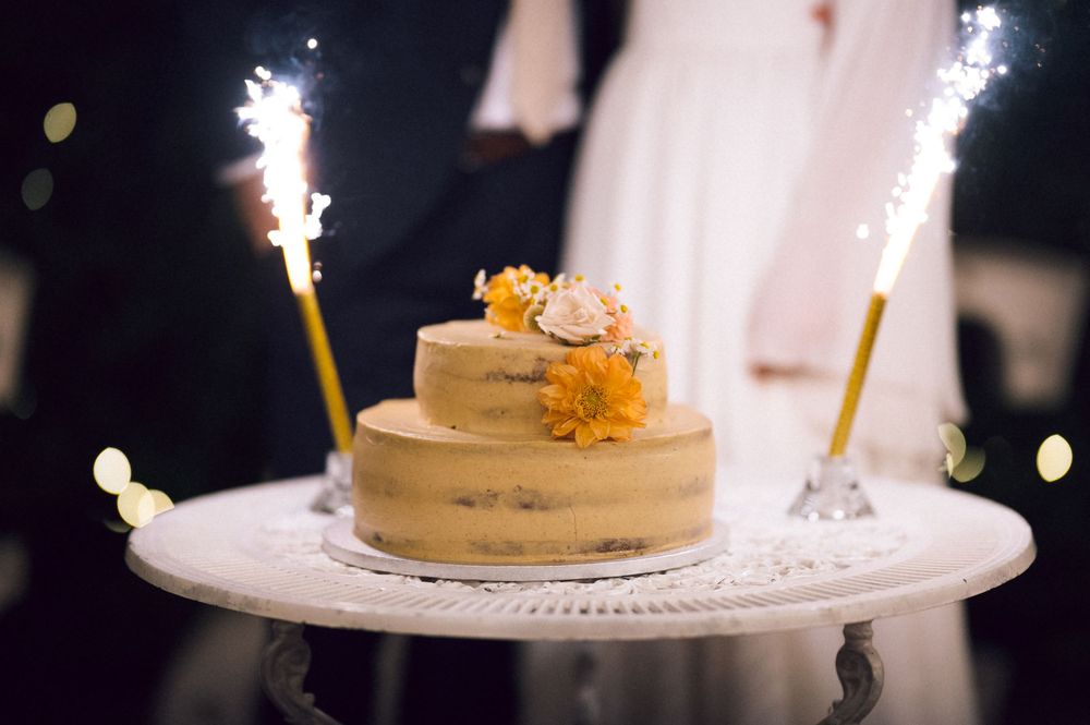 Two-tier naked wedding cake with orange flowers and sparklers on white stand.