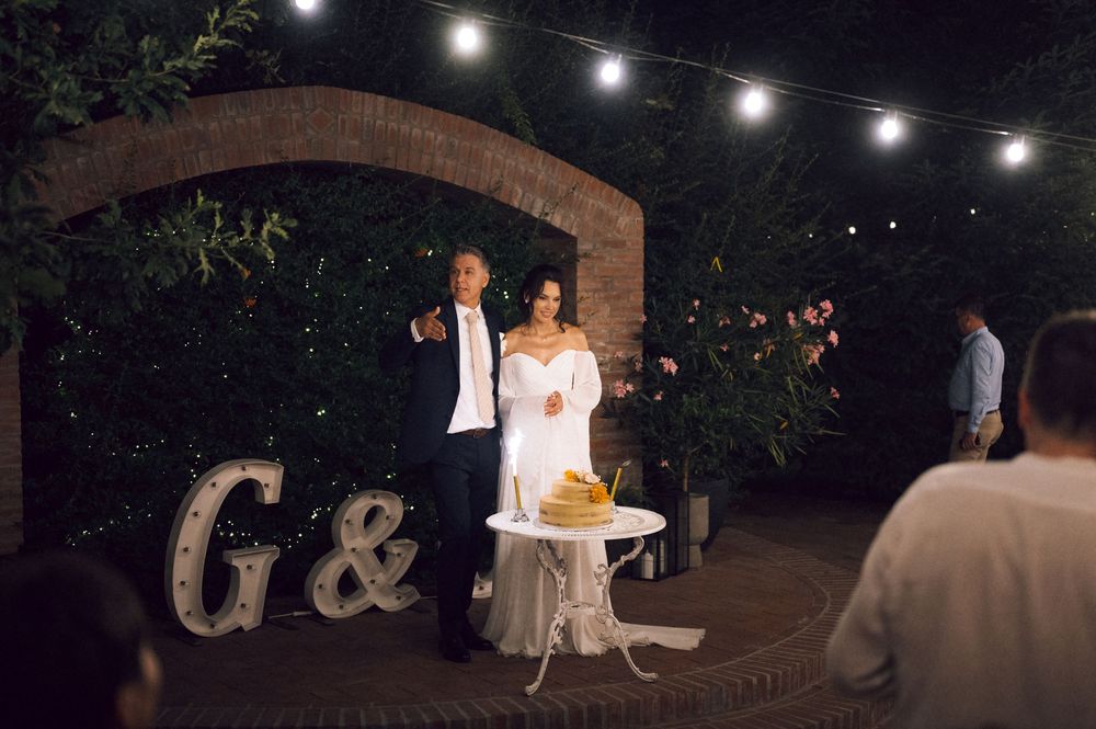 Bride and groom cutting wedding cake outdoors at night under string lights with marquee letter decorations.