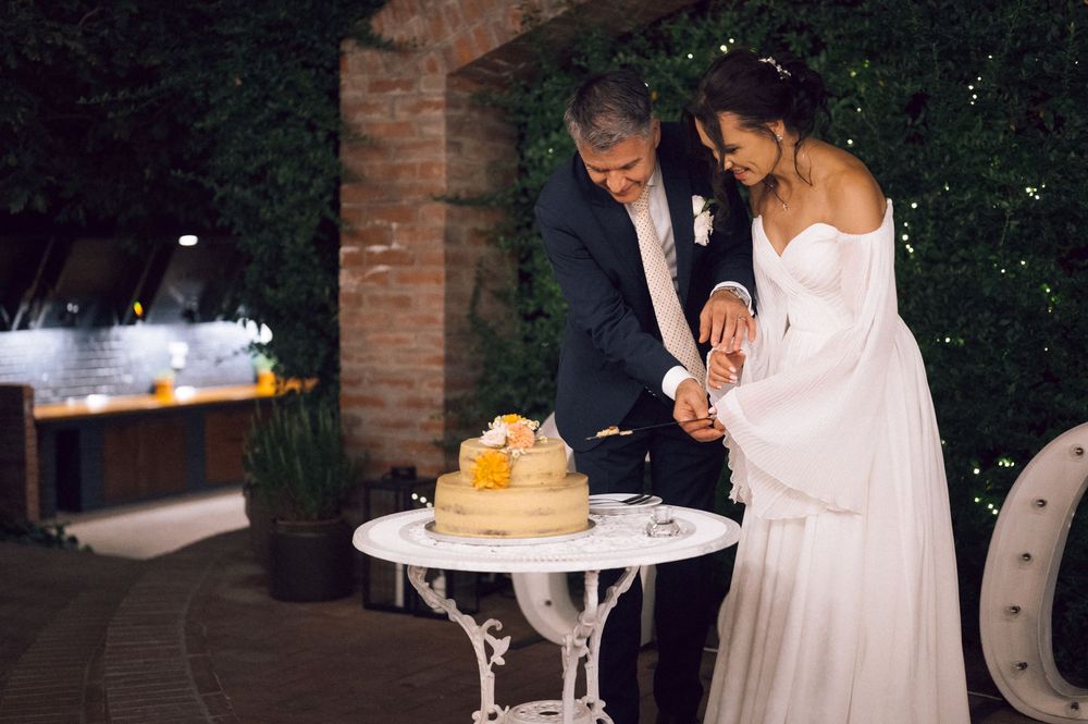 Bride and groom cutting wedding cake outdoors at night, elegant white dress, suit, decorated table, lush greenery backdrop.