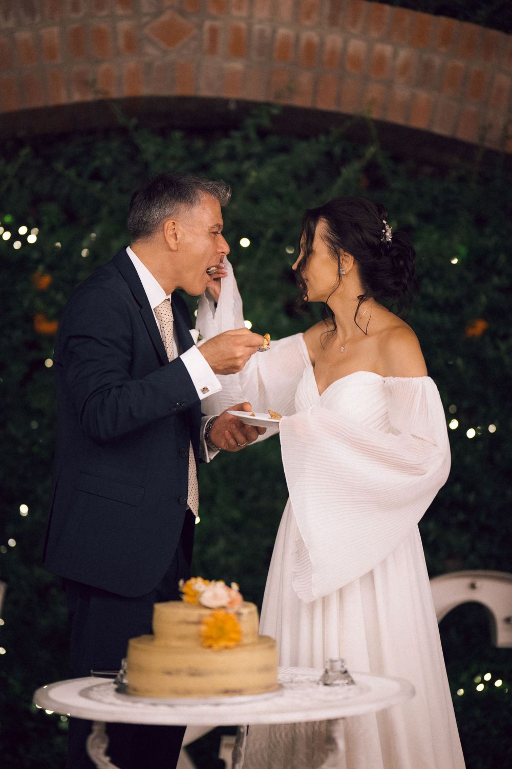 Bride and groom cutting wedding cake outdoors at night, sharing a sweet moment under a brick arch with twinkling lights.