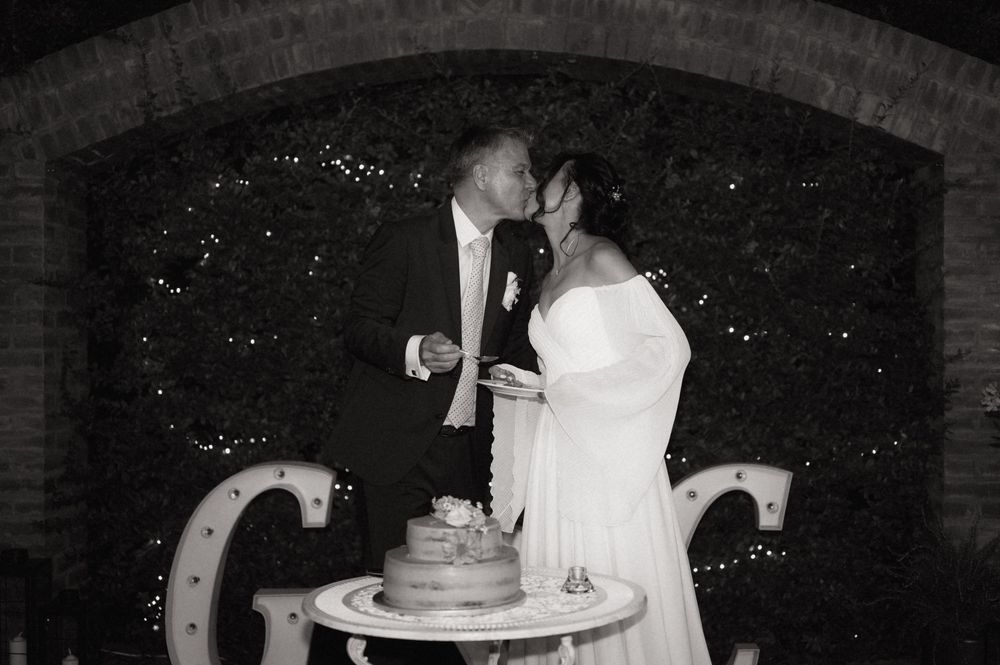 Black and white photo of a bride and groom kissing while cutting their wedding cake outdoors at night.