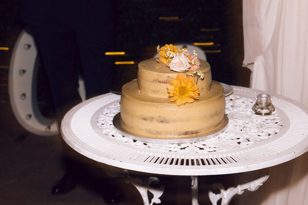 Two-tier naked wedding cake with yellow and pink flowers on a white ornate table.