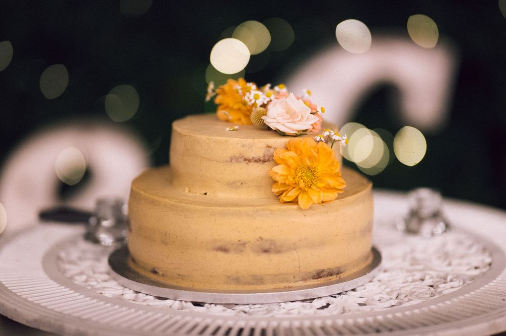 Two-tier naked wedding cake decorated with yellow marigolds and white flowers on a white doily cake stand.