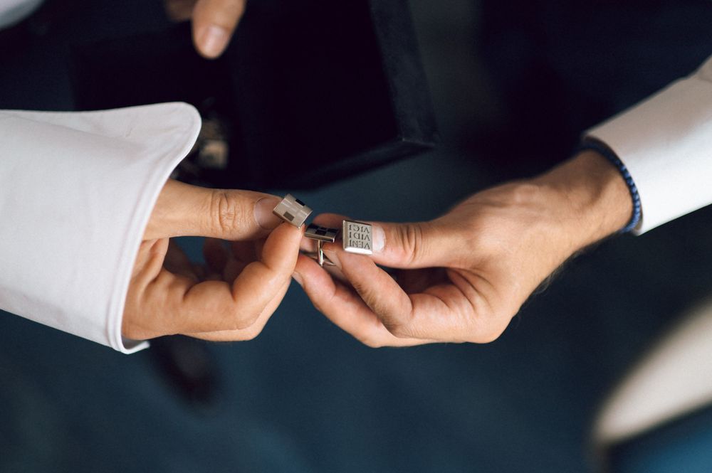 Man fastening a decorative silver cufflink on a white dress shirt sleeve while wearing a dark suit jacket.