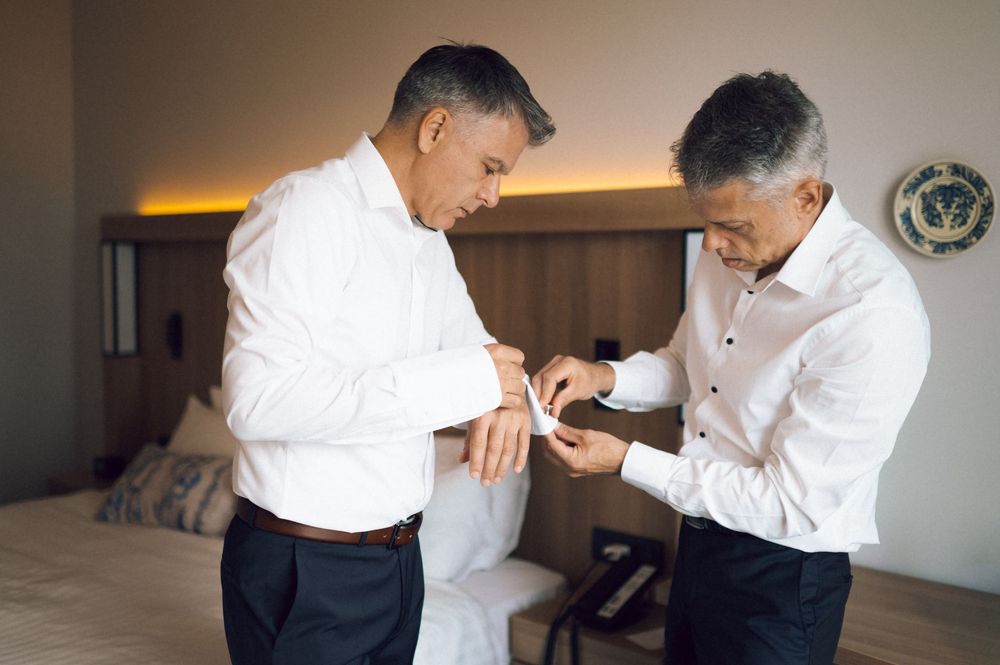 Two men in white dress shirts adjusting cufflinks in a hotel room before a formal event.