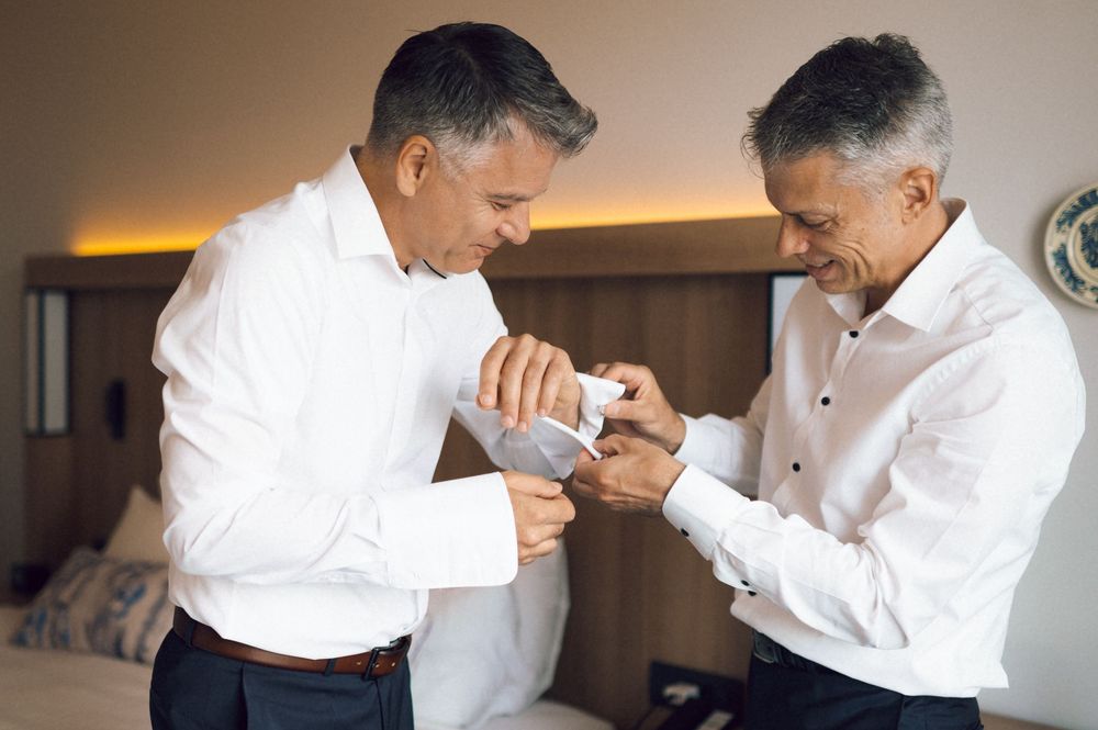 Two men in white dress shirts helping each other with cufflinks in a hotel room before a formal event.