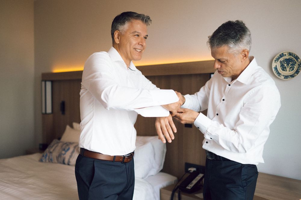 Two mature men in white shirts adjusting cufflinks in a hotel room, smiling and getting dressed together.