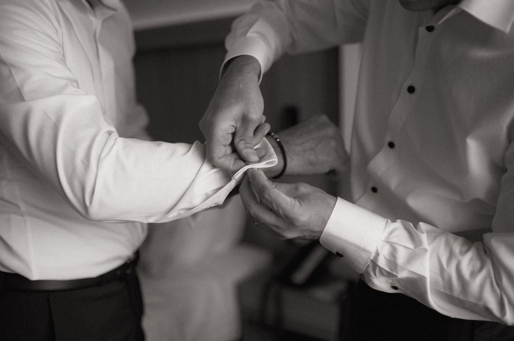 Black and white photo of two men in dress shirts fastening cufflinks, preparing for a formal wedding event.