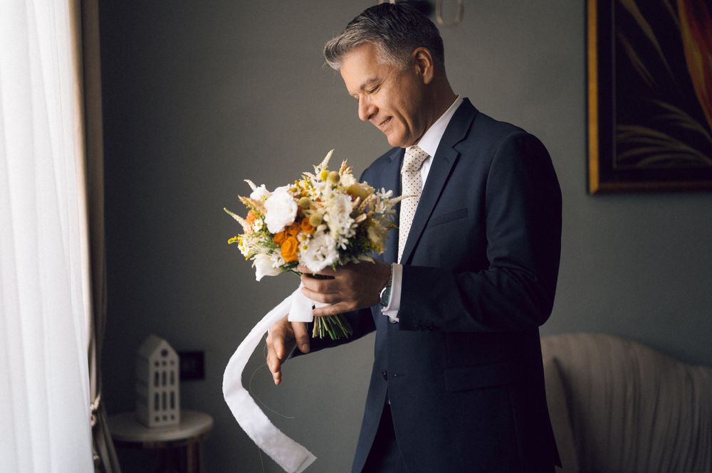 Groom in dark suit holds a white and orange wedding bouquet with ribbon indoors.