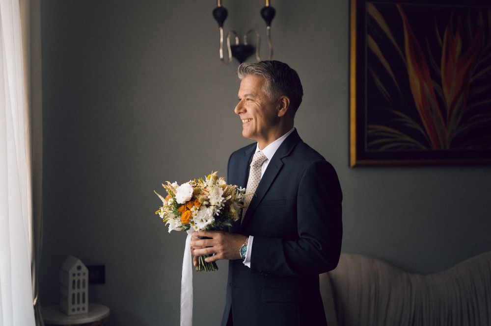 Smiling groom in navy suit holding a colorful bridal bouquet by a window in a softly lit room.