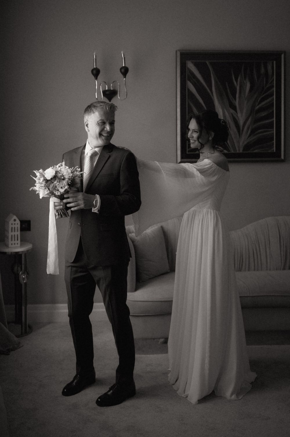 Black and white photo of a groom holding bouquet while bride adjusts veil in elegant room.