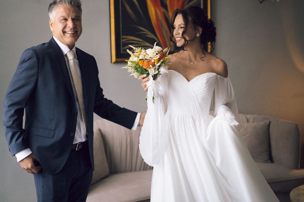 A bride in a white off-shoulder gown holds a bouquet beside a smiling man in a navy suit indoors.