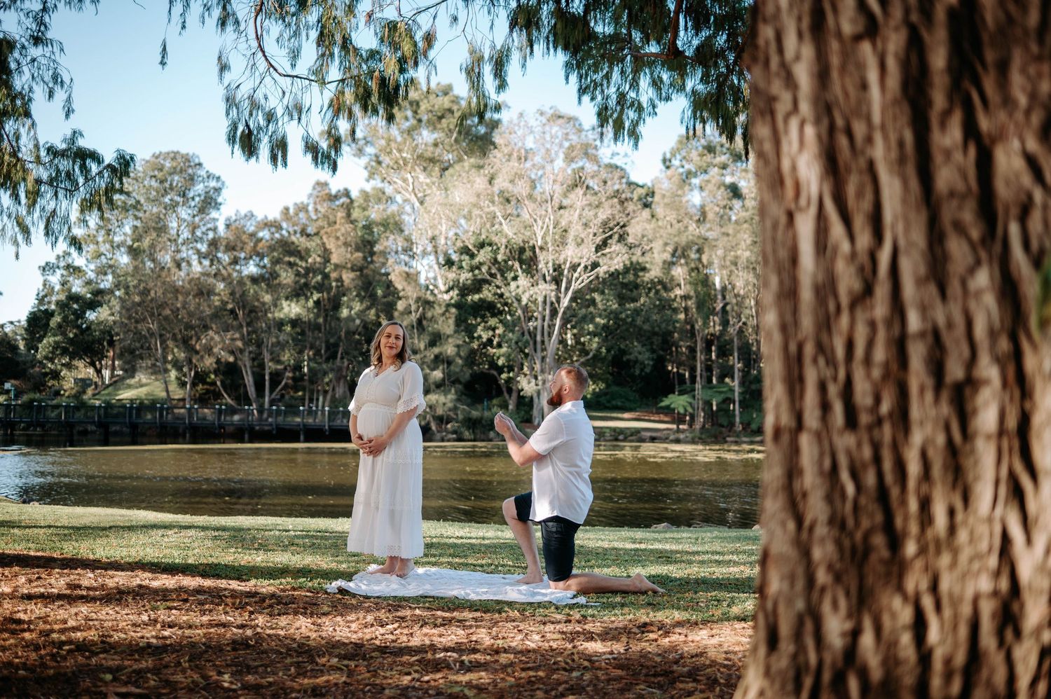 Couple practicing yoga together on a mat by a tranquil river in a lush Australian park setting.