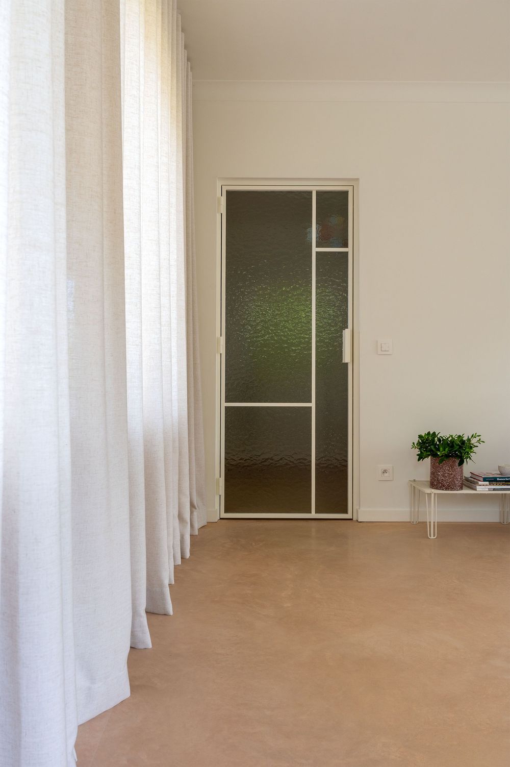 Minimalist room with sheer white curtains, textured glass door, beige floor, and small plant on white side table.