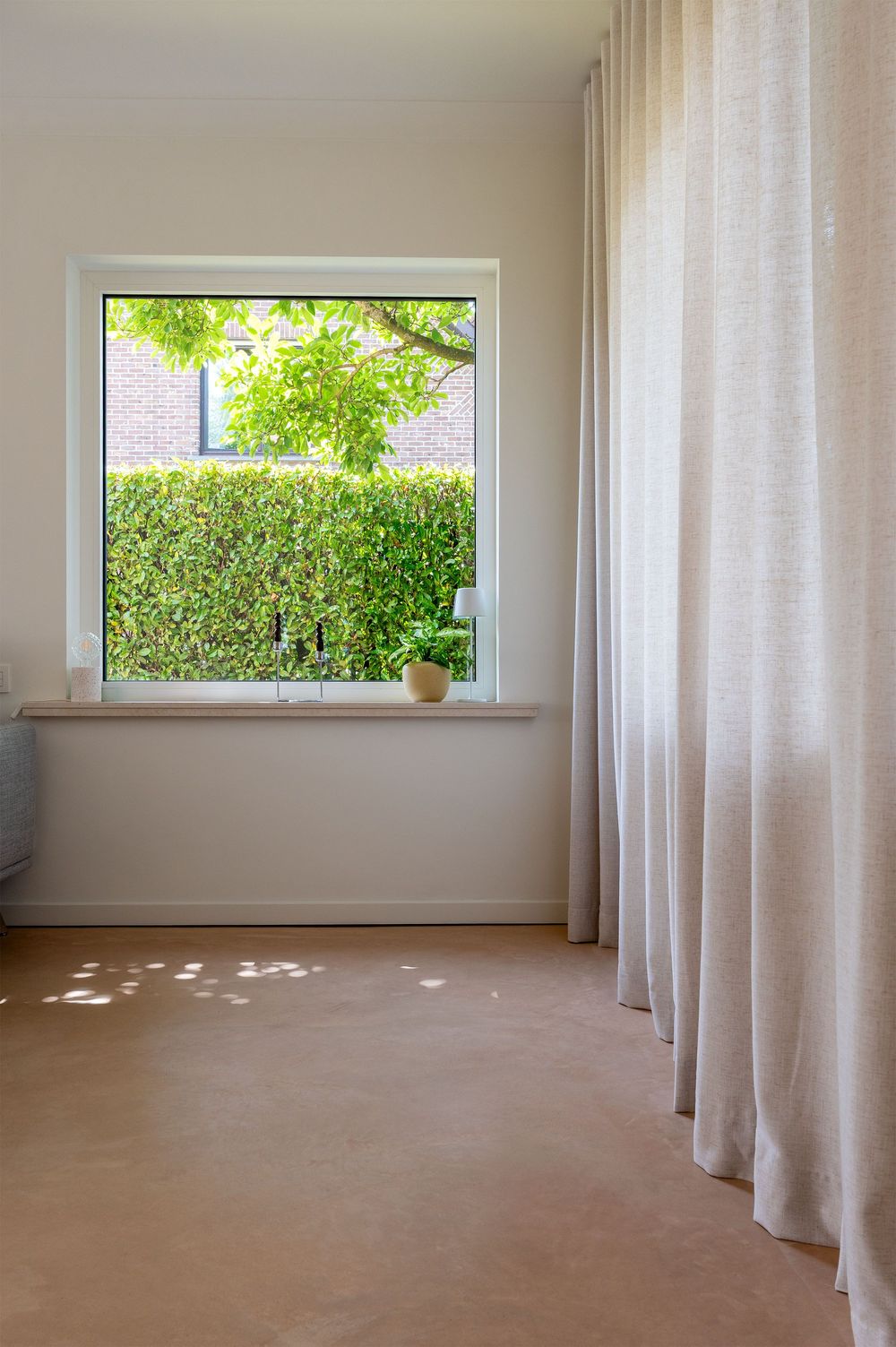 Bright minimalist room with sheer white curtains, a small potted plant on the windowsill, and a view of green hedges outside.