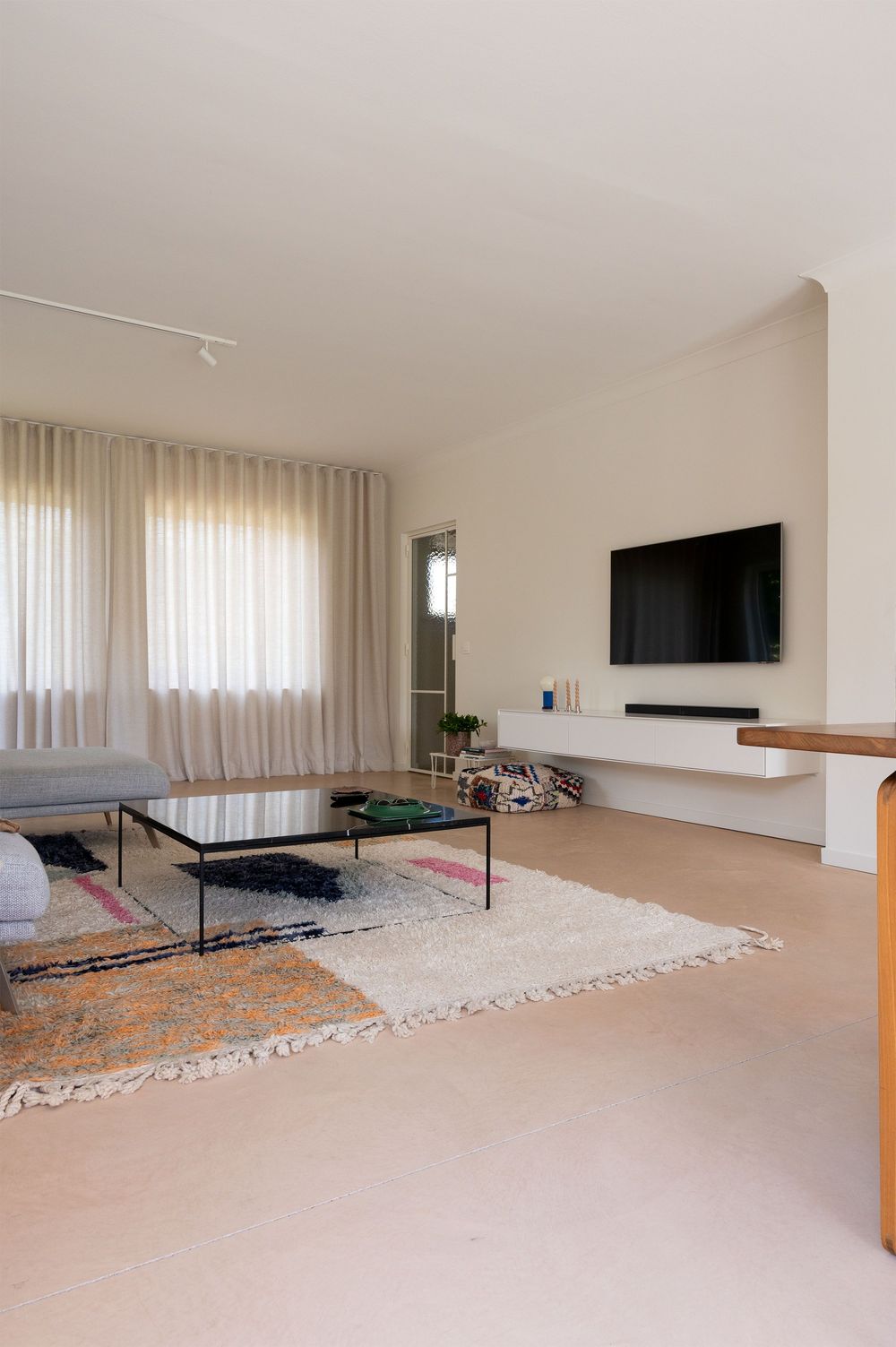 Modern minimalist living room with wall-mounted TV, sheer curtains, layered rugs, and a glass coffee table on light flooring.