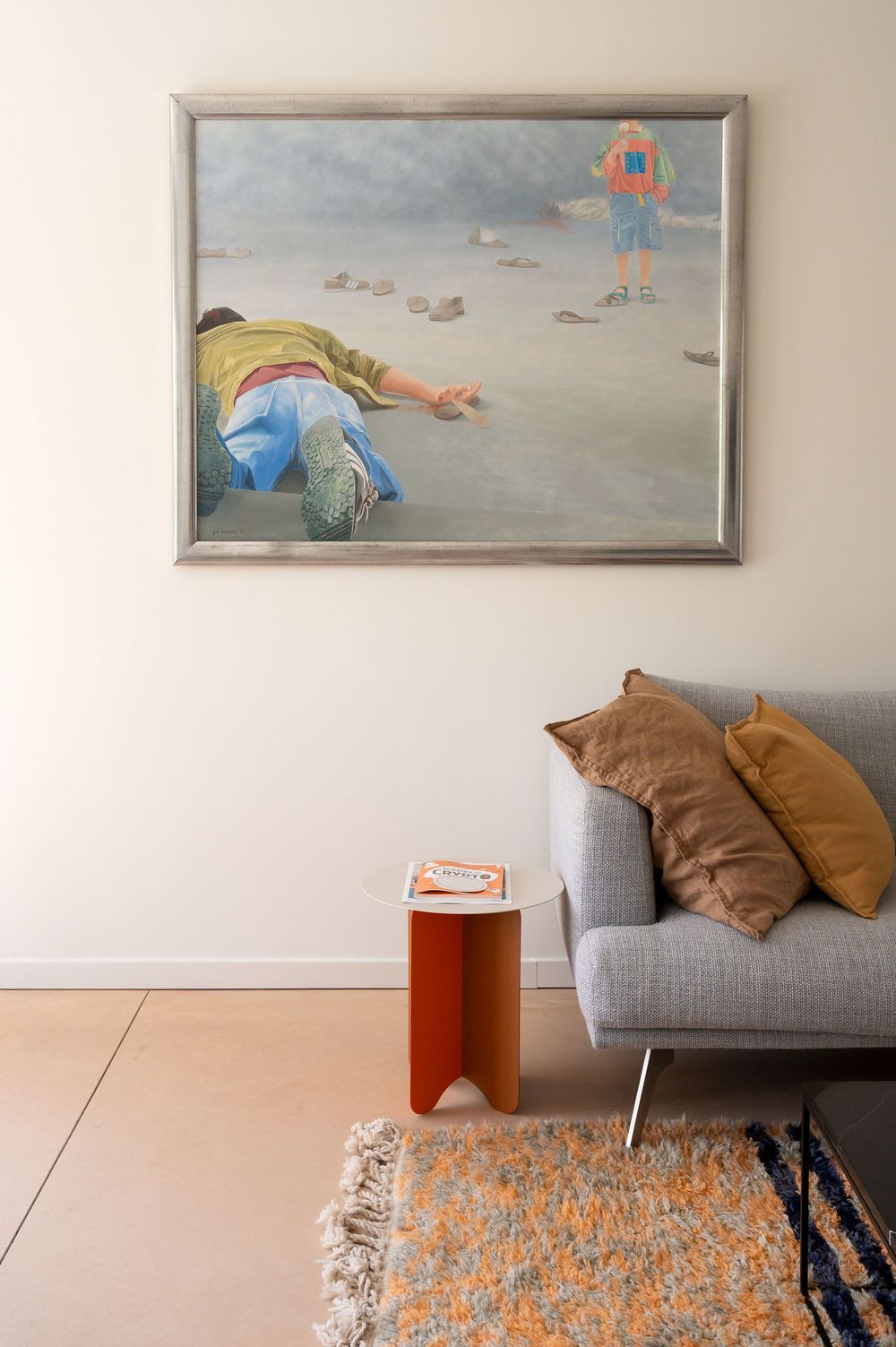 Modern living room with gray sofa, orange side table, patterned rug, and large framed painting of figures on a beach.
