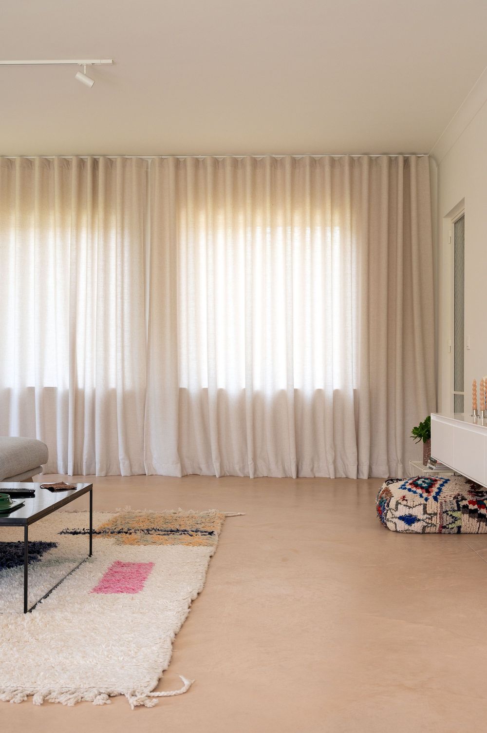 Minimalist living room with sheer white curtains, layered rugs, and a low black coffee table on light wood floors.