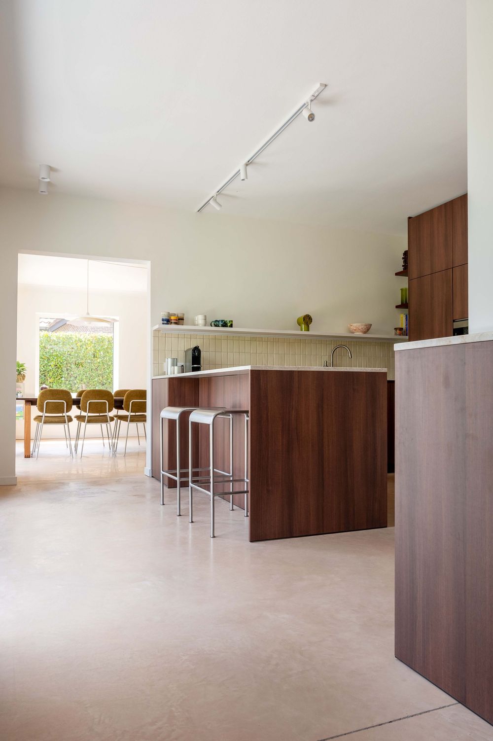Modern kitchen with walnut island, bar stools, open shelving, and dining area with yellow chairs visible through window.