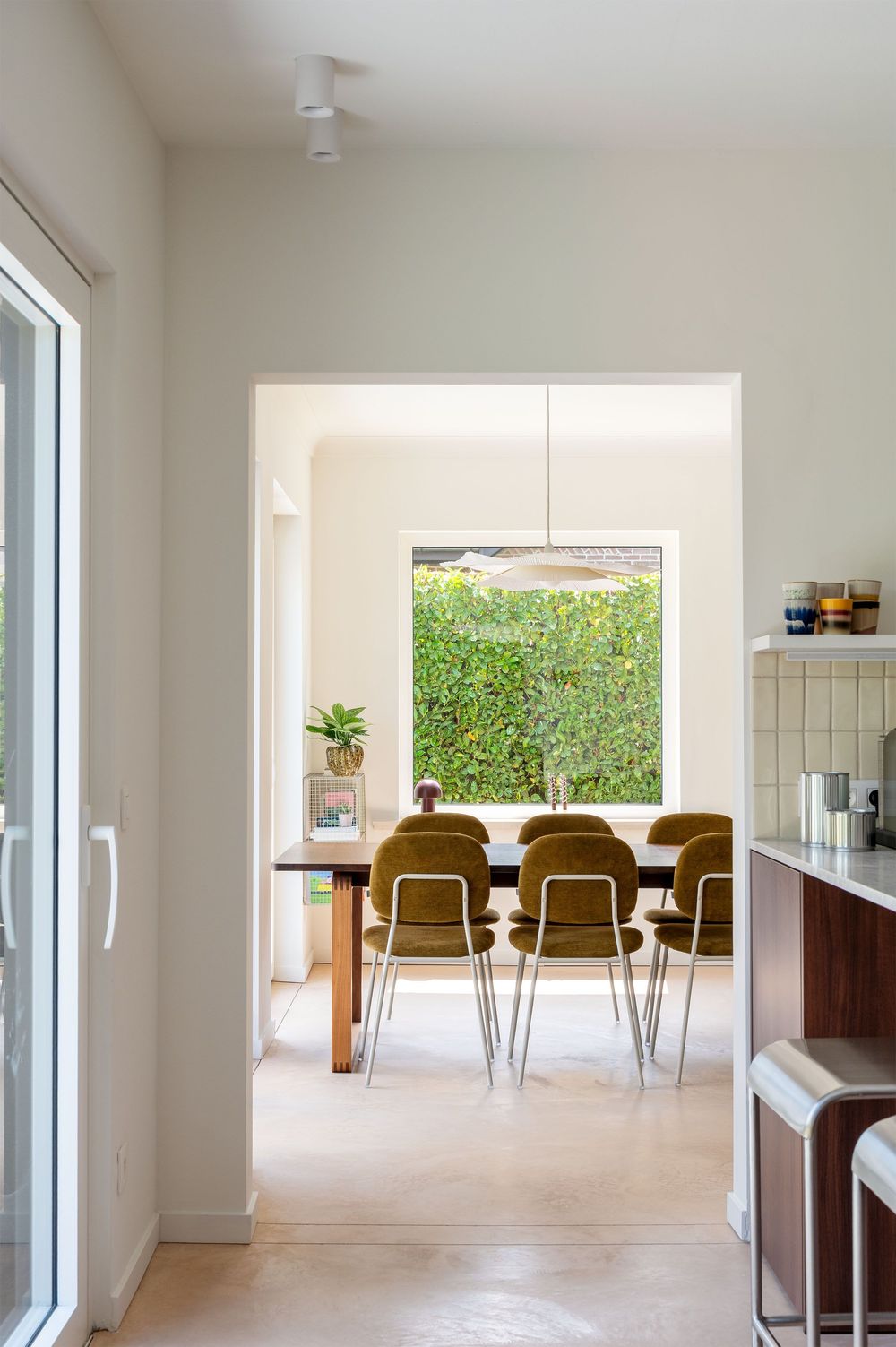 Modern dining room with olive green chairs, wooden table, and lush green garden view through large window.