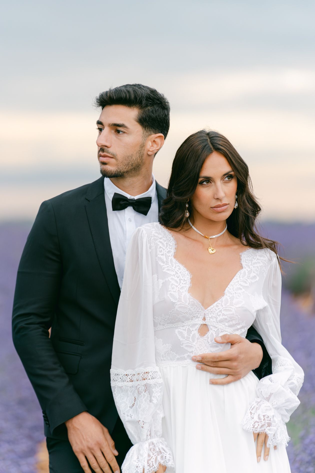 Elegant couple in lavender field, groom in black tuxedo embracing bride in white lace boho wedding dress.