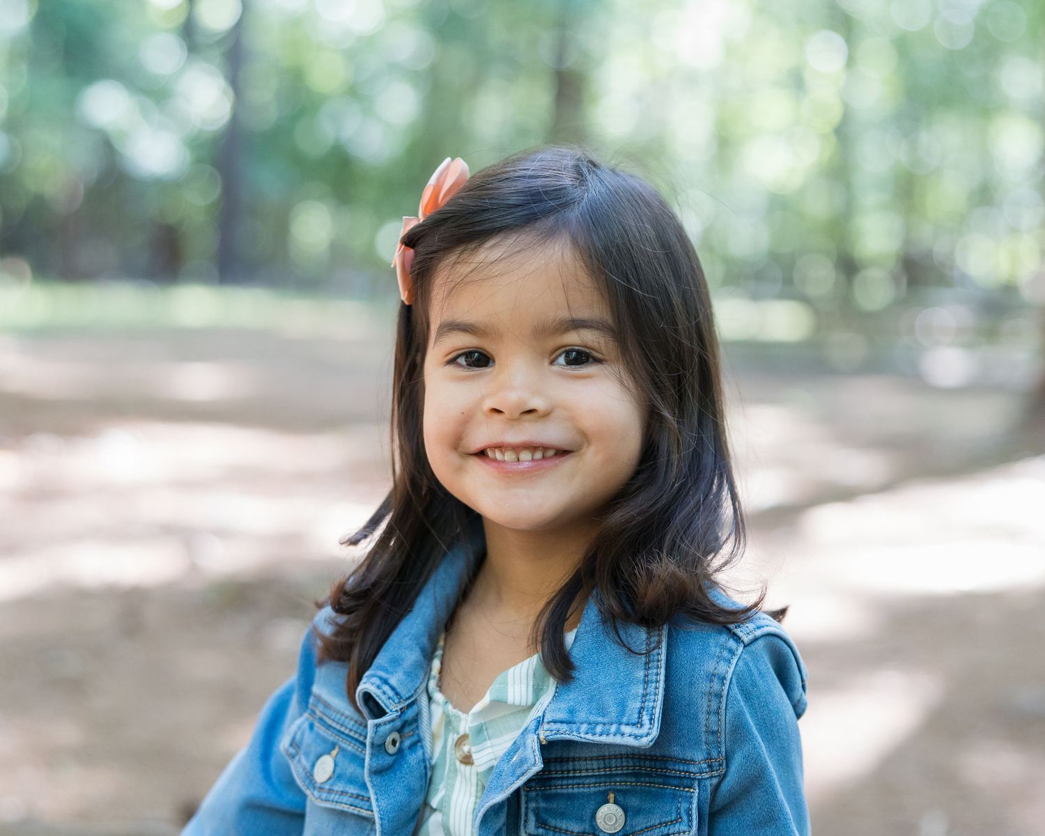 Smiling toddler girl with dark hair and hair clip wearing denim jacket outdoors in a sunlit park.