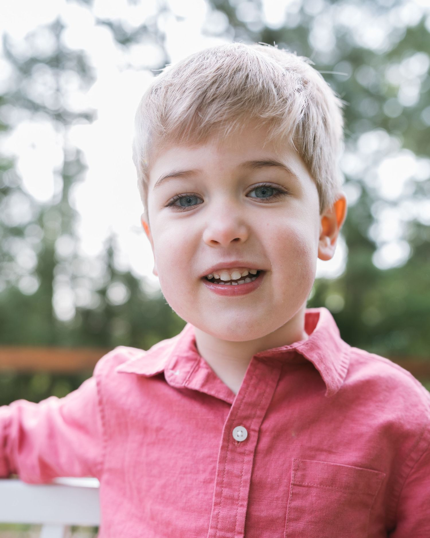 A young boy with blonde hair and blue eyes smiles wearing a pink button-up shirt outdoors.