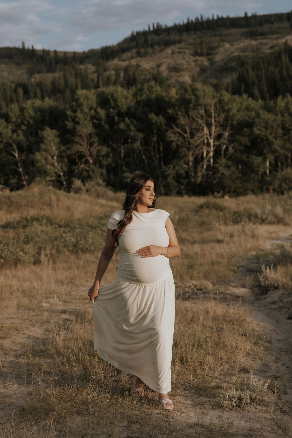 Pregnant woman in white maxi dress standing in golden field with forested hills backdrop during maternity photoshoot.