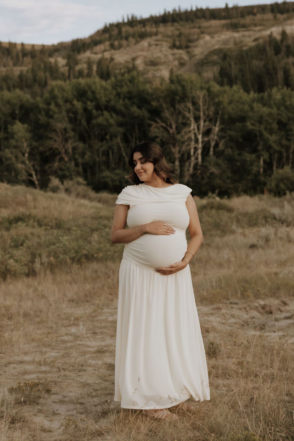 Pregnant woman in white dress cradling pregnant bump in Alberta's badlands during a maternity photoshoot