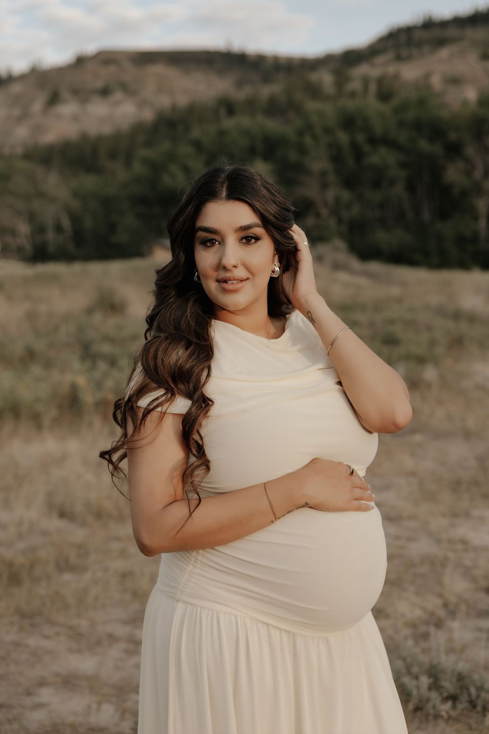 Pregnant woman in white dress posing outdoors in a field with green hills in the background.