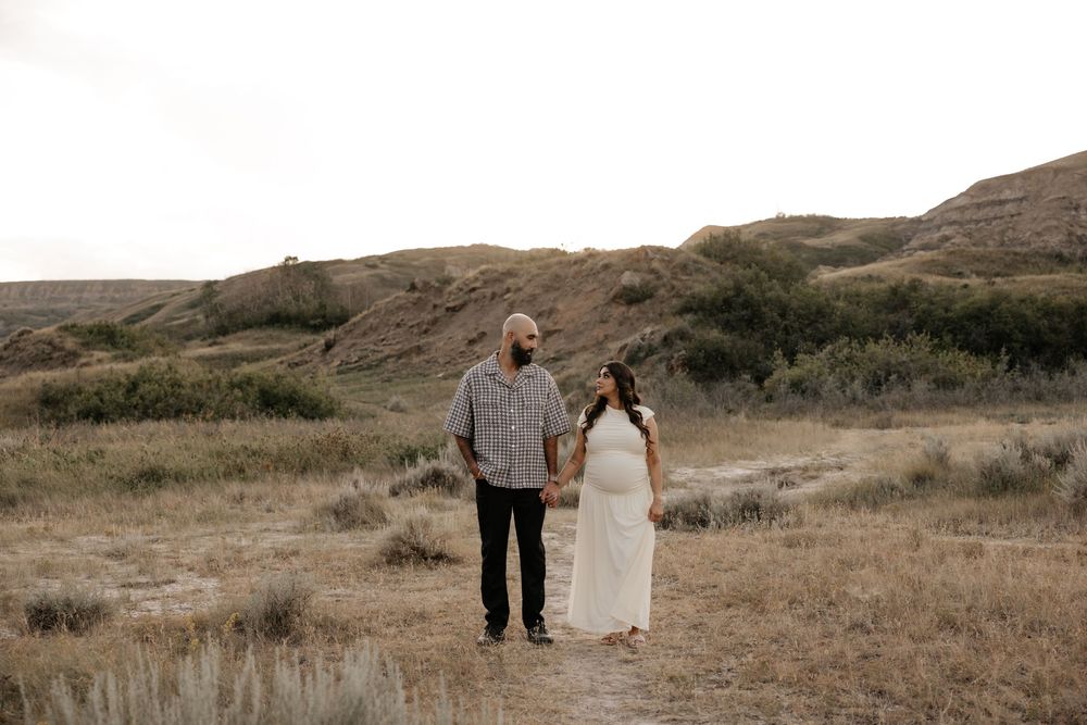 Couple holding hands on a dirt path in a golden field with rolling hills and shrubs in the background.