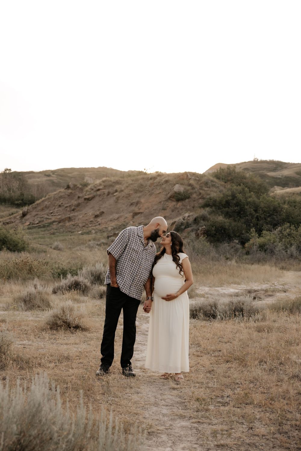 Couple sharing a tender kiss during maternity photo shoot in golden desert landscape, woman in white dress cradling baby bump.